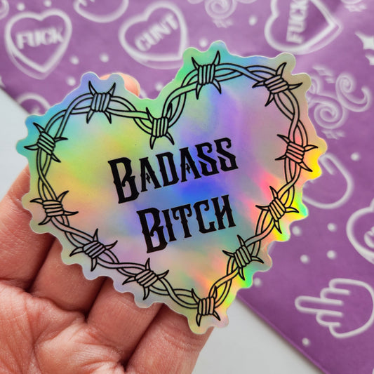 Holographic sticker with 'BADASS BITCH' text held by a hand against a purple background with white designs.